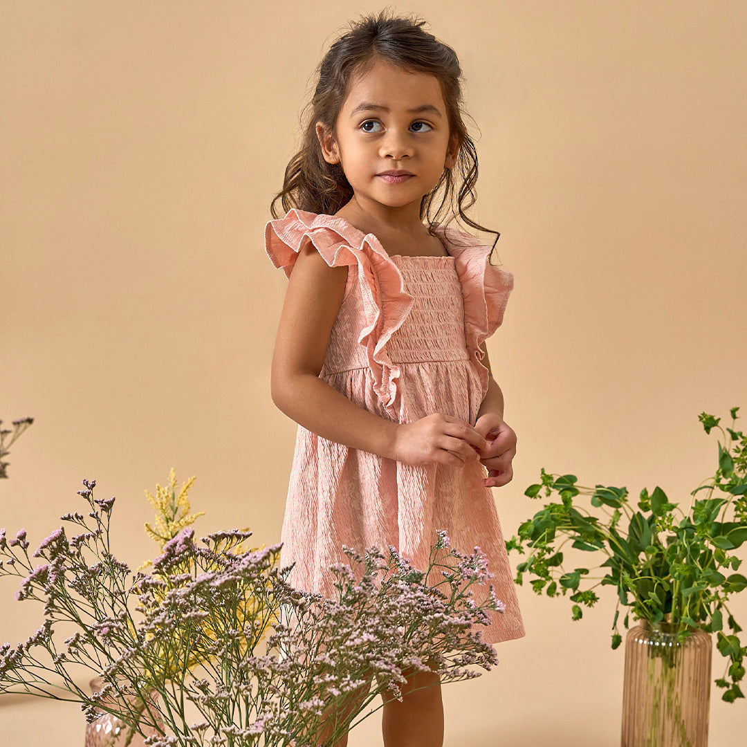 Little girl in a ruffled pink dress standing among flowers. Click here to browse playful dresses from Modern Moments.